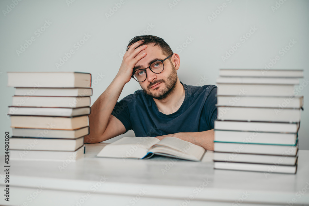Young white European brunette man nerd in glasses teaches lessons, reads books, studied at university, prepares for exams, has many books at home in white backround table. Young white European brunette man nerd in glasses teaches lessons, reads books, studied at university, prepares for exams, has many books at home in white backround table.