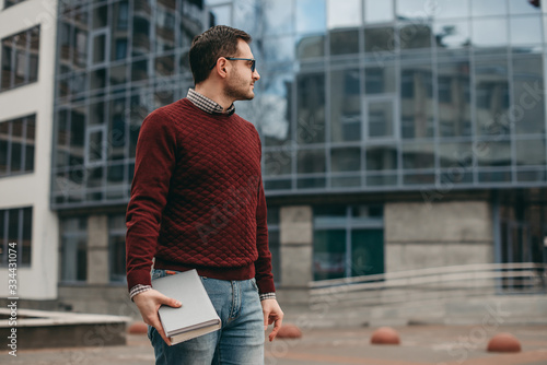 Portrait of a man in glasses for vision wearing a shirt and sweater, with , frame, with books, notebook in his hands, on the background of a modern building. Education, institute, business, outside