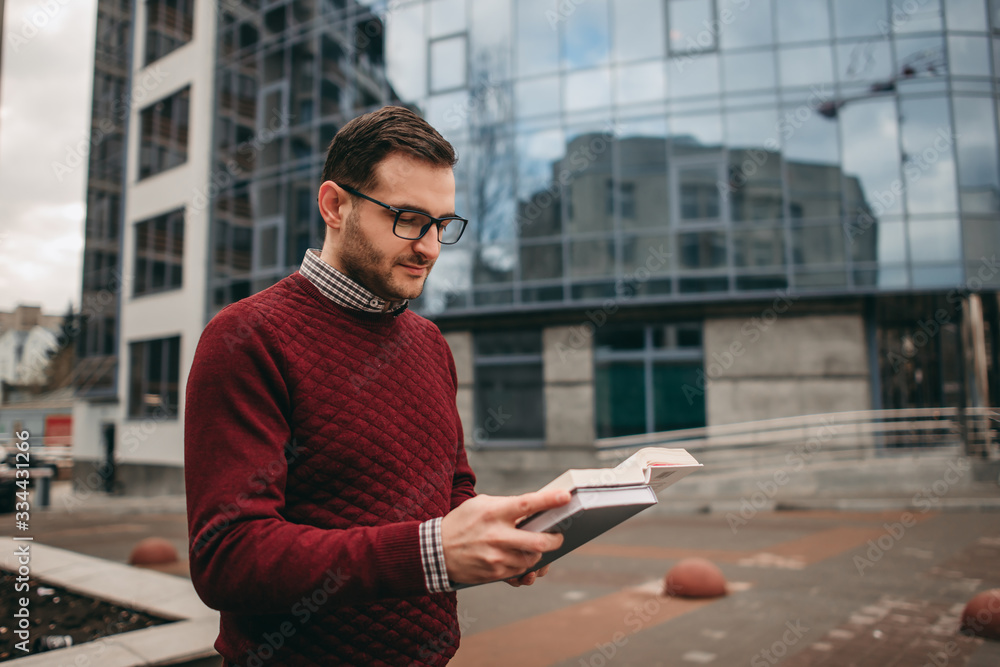 man wearing glasses wearing a shirt and sweater, wearing glasses for vision, reading a book, a notebook, against the background of a modern building. Education, institute, business man wearing glasses wearing a shirt and sweater, wearing glasses for vision, reading a book, a notebook, against the background of a modern building. Education, institute, business