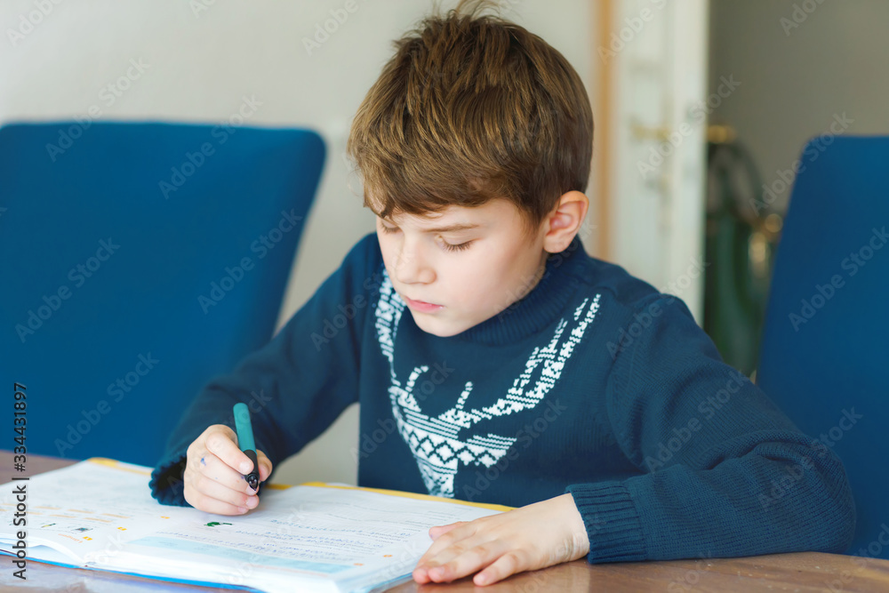 Hard-working happy school kid boy making homework during quarantine time from corona pandemic disease. Healthy child writing with pen, staying at home. Homeschooling concept