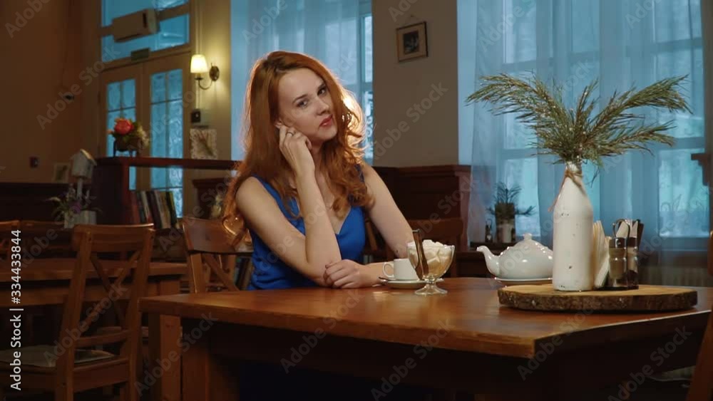 The beautiful sexy girl with red hair in the blue dress sitting in cafe, posing and smiling. Slowmo. The beautiful sexy girl with red hair in the blue dress sitting in cafe, posing and smiling. Slowmo.