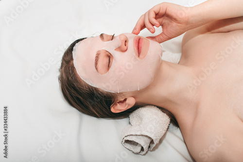 Portrait of young beautiful european woman lying in beauty salon with collagen silicone fabric with white mask on face on white background. Beauty treatments, the secret of youth, health.