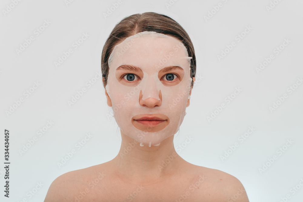 Fototapeta premium Portrait of young beautiful european woman with blue eyes with collagen silicone fabric with white mask on face on white background. Beauty treatments, the secret of youth, beauty, health.