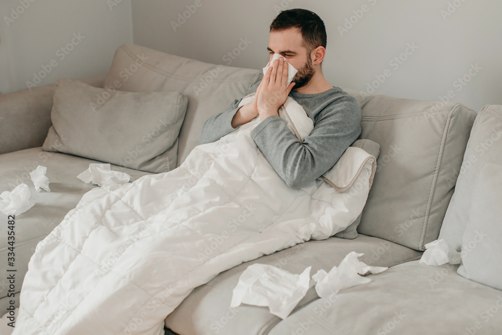 Young sick bearded european man sneezes into napkin at home on gray sofa with white blanket. Disease, protection, coronavirus, virus, disease, flu, respiratory dressing. Young sick bearded european man sneezes into napkin at home on gray sofa with white blanket. Disease, protection, coronavirus, virus, disease, flu, respiratory dressing.