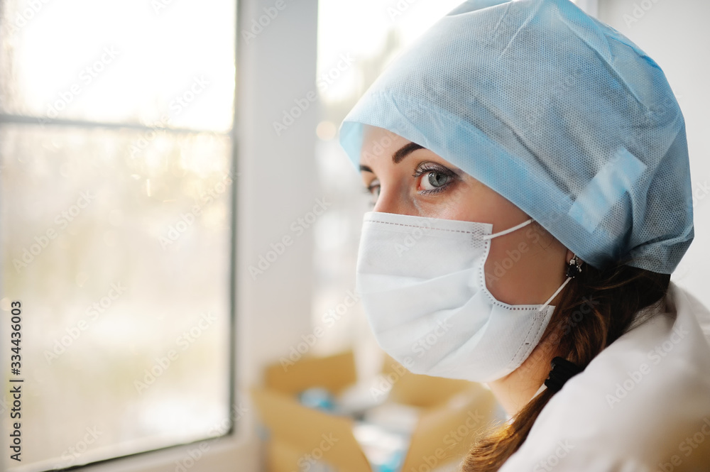 portrait of a young beautiful female doctor in a medical cap and sterile mask on the background of a rainy window and clinic. Medical concept. Copy space portrait of a young beautiful female doctor in a medical cap and sterile mask on the background of a rainy window and clinic. Medical concept. Copy space