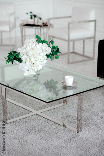 Fragment of a luxurious interior of a modern apartment. White wall, glass table, composition of white flowers and a navy sofa.