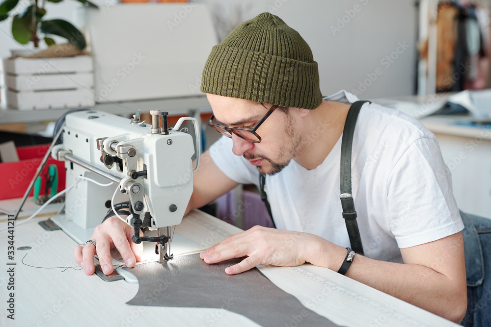 Serious young male leatherworker bending over electric sewing machine