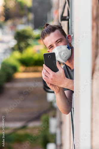 Young man in a medical mask looks out of the apartment window and photographs a neighbor using a mobile phone. Quarantined during the coronavirus epidemic