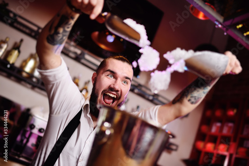 a handsome bearded bartender in a white shirt pours ice for cocktails and screams happily against the bar at a nightclub party