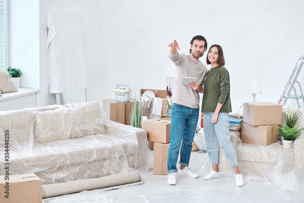 Young man pointing at the corner of living-room with his wife standing near by Young man pointing at the corner of living-room with his wife standing near by