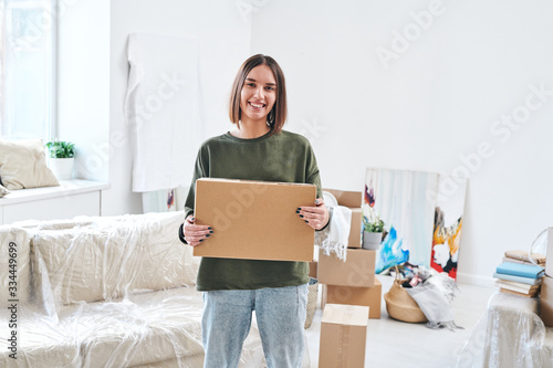 Young smiling woman holding carton box while standing in living-room