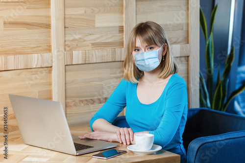 Portrait of young blonde woman with surgical medical mask in blue t-shirt is sitting and working on laptop and looking at camera. Indoor working, medicine and health care concept.