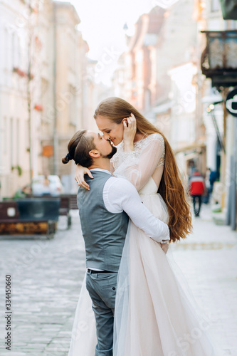 Handsome groom holds his bride in hands kissing on lips against morning street background © Yuliia