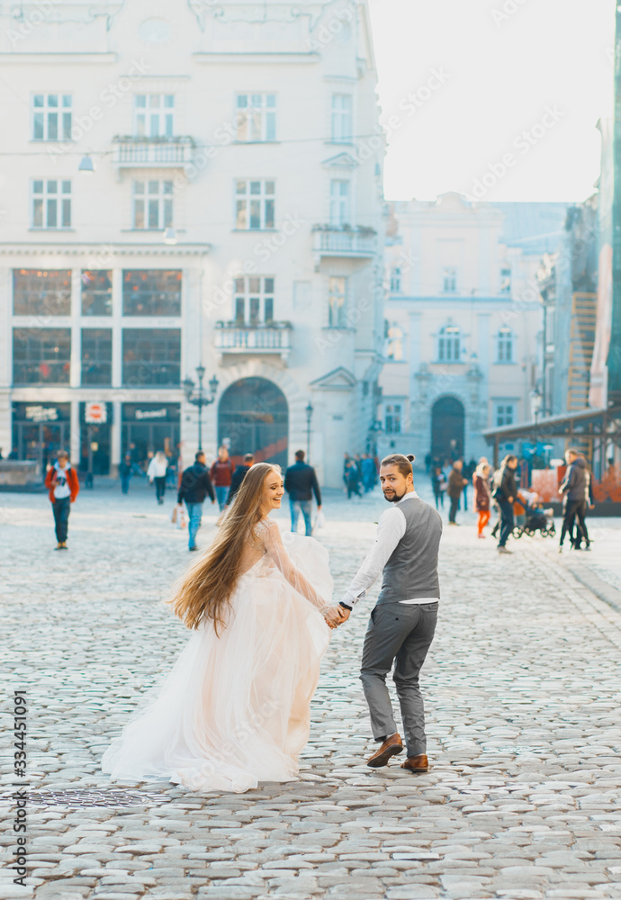 Incredible young couple running against the backdrop of morning city looking back. Lovely bride with long hair. Handsome groom in gray suit..