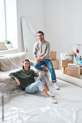 Young amorous couple in casualwear looking at you while relaxing by new couch