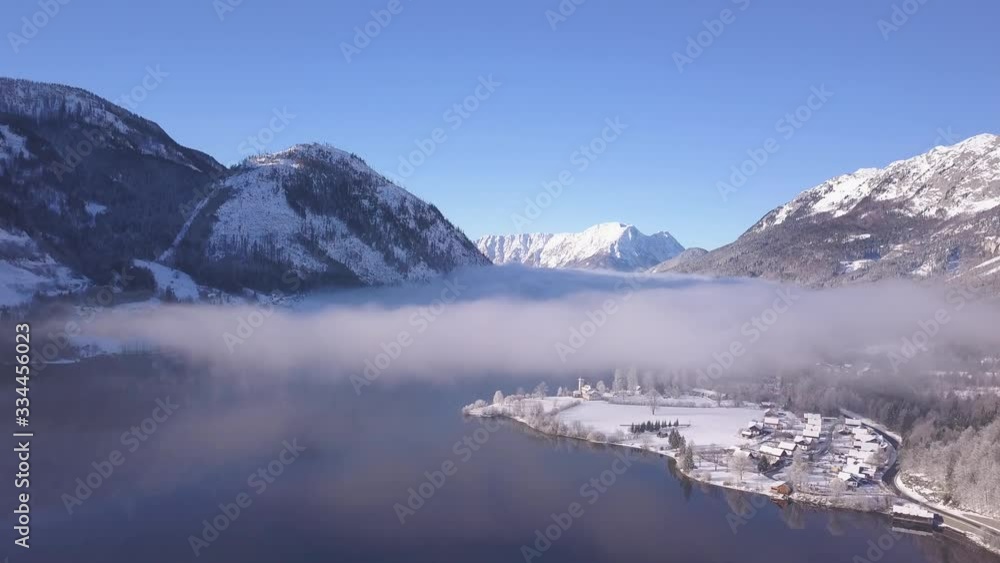 High aerial, fog over lake in snow covered mountainous valley with small village High aerial, fog over lake in snow covered mountainous valley with small village
