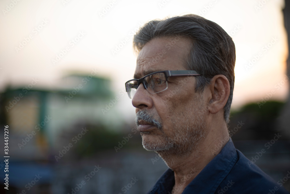 Naklejka premium Portrait of an An old/aged Indian Bengali man in blue shirt is standing on a rooftop under the open sky in front of a urban landscape. Indian lifestyle