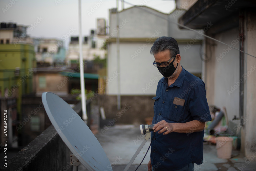 Portrait of an Indian old man wearing corona preventive mask repairing dish antenna in home isolation. Indian lifestyle, disease and home quarantine.