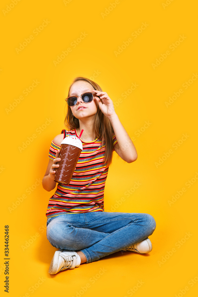 Fototapeta premium Cute caucasian young girl in t-shirt and sunglasses drinking milk cocktail on yellow background
