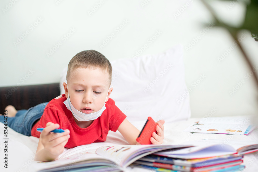 A boy lying on the couch doing lessons on home quarantine using his phone and books. A boy lying on the couch doing lessons on home quarantine using his phone and books.