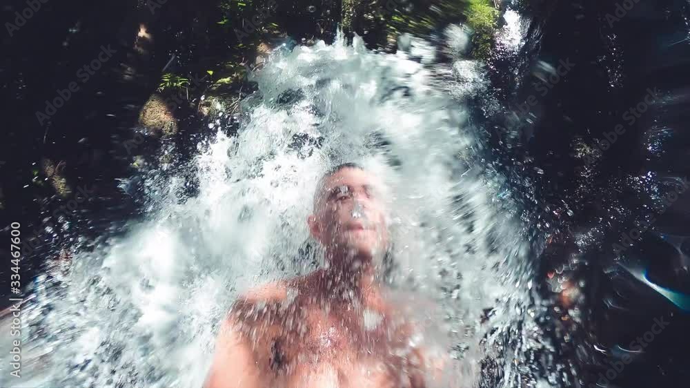 Man refreshing under a beautiful waterfall, slow motion, upward view Man refreshing under a beautiful waterfall, slow motion, upward view