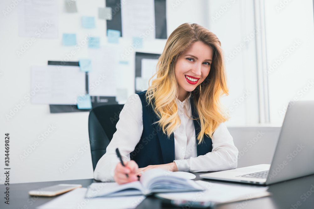 young blond smiling businesswoman works in her laptop in modern office, taking notes to her business planner,looks confident and busy, multitasking, work concept young blond smiling businesswoman works in her laptop in modern office, taking notes to her business planner,looks confident and busy, multitasking, work concept