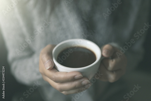 Closeup of female hands with a cup of hot coffee. in the morning soft sunlight. Beautiful girl in grey sweater.