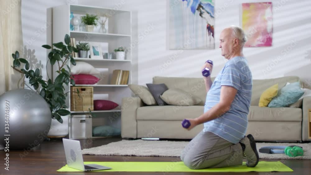 Tracking shot of senior man standing on knees on mat in the living room, watching video workout on laptop and doing dumbbell curls while training at home Tracking shot of senior man standing on knees on mat in the living room, watching video workout on laptop and doing dumbbell curls while training at home