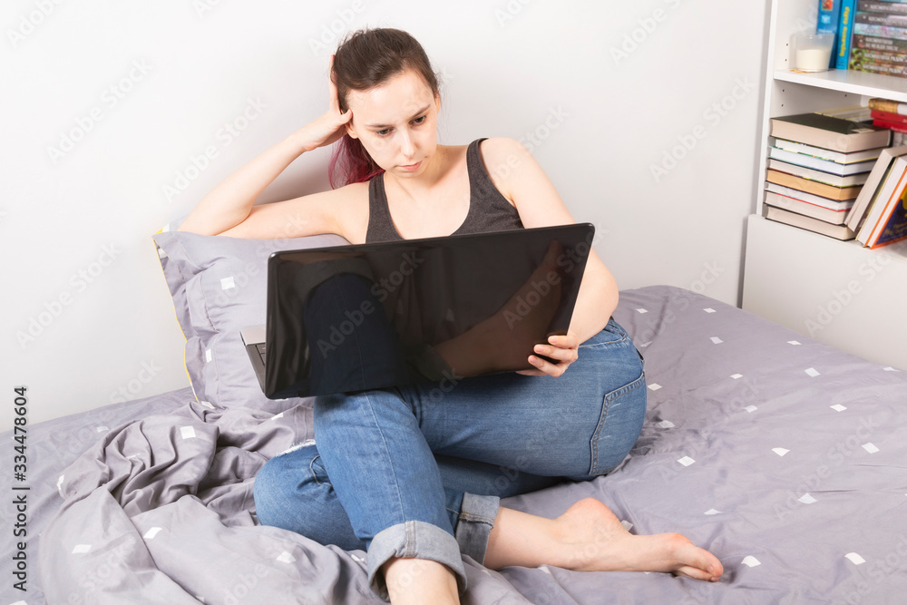 Concept - work from home, self-isolation. Young girl lies in bed looking into laptop Concept - work from home, self-isolation. Young girl lies in bed looking into laptop