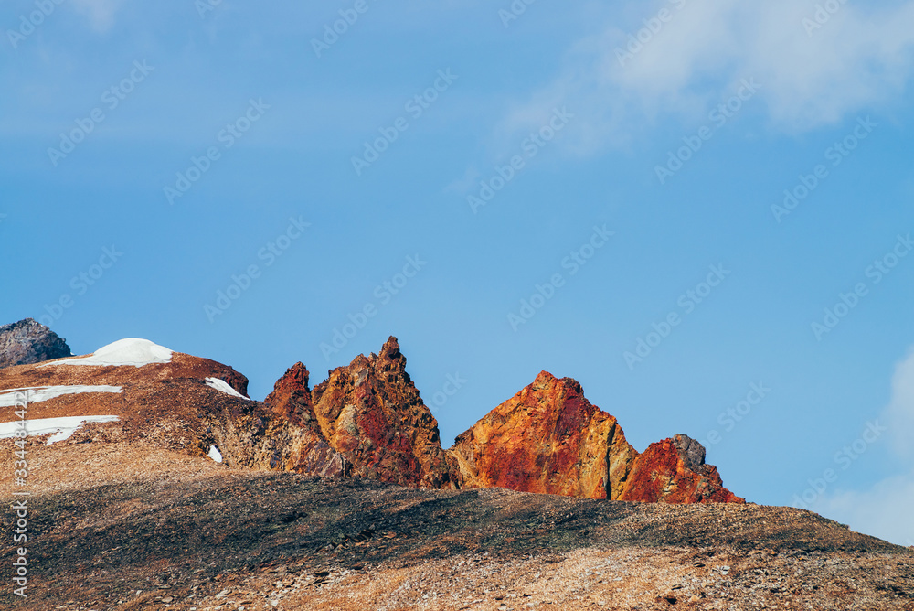 Obraz premium Awesome sunny mountain with red orange brown crags in top under blue sky. Colorful stony hill with snow in sunshine. Wonderful alpine scenery with rock in red color. Beautiful vivid highland landscape