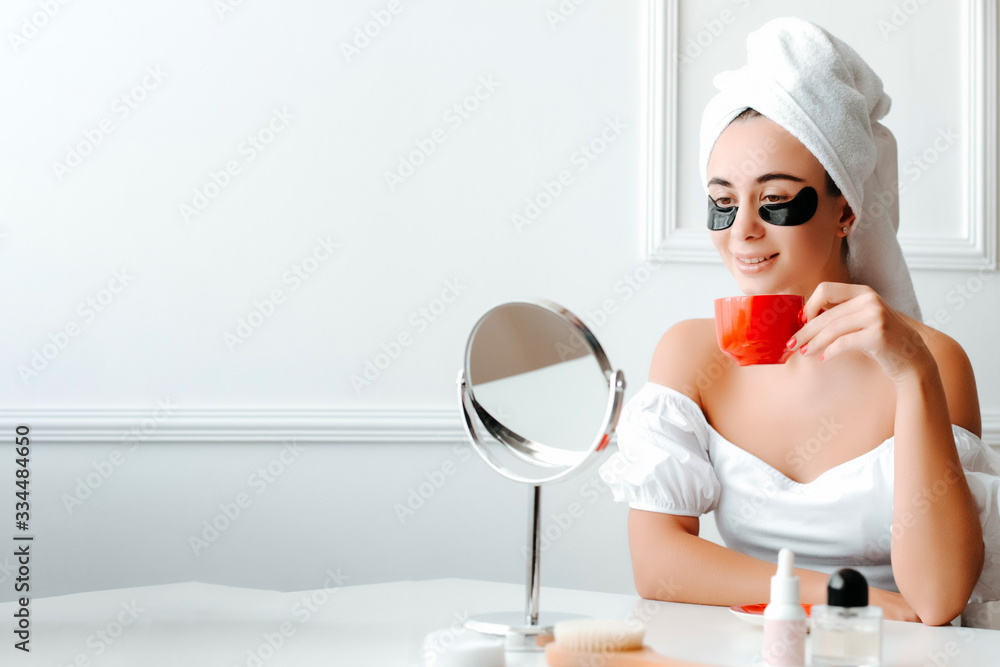 Beautiful smiling girl near the mirror, sitting at the table, drinking morning coffee. The girl with patches under the eyes, beauty routine. Beautiful smiling girl near the mirror, sitting at the table, drinking morning coffee. The girl with patches under the eyes, beauty routine.