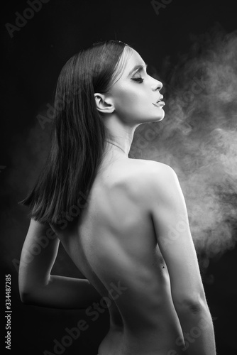 A beautiful slim topless girl with avant-garde makeup sensually poses covering her breast with her hands, in smoke on a black background.  Artistic, fashionable, advertising monochrome design.