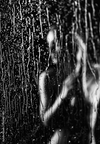 A beautiful slim nude girl in defocus sensually poses behind a window glass, on which streams and drops of rain water flow down on a black background. Artistic blurred monochrome design.