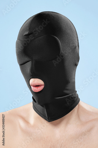 A young man with naked shoulders is wearing a black elastic blindfold mask with a mouth hole. The man in the breathable mask is posing on the blue backdrop.