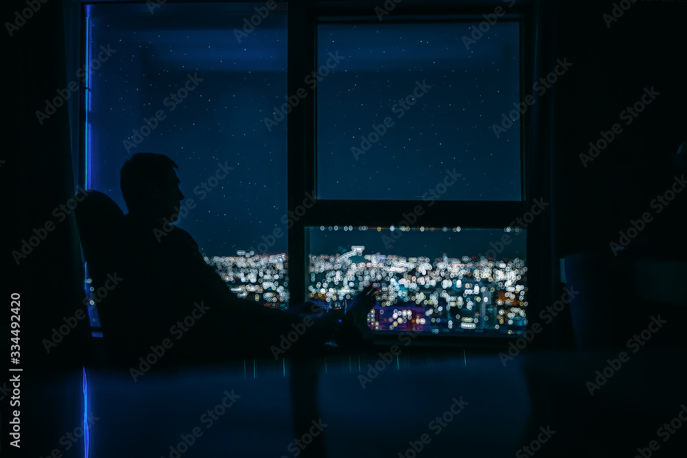 Silhouette of a young man on the panoramic window background with the glass of wine in his hand and is looking on the night city landscape with stars on the sky Silhouette of a young man on the panoramic window background with the glass of wine in his hand and is looking on the night city landscape with stars on the sky
