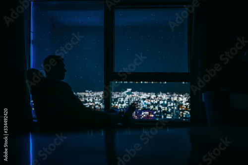 Silhouette of a young man on the panoramic window background with the glass of vine in her hand and is looking on the night city landscape with stars on the sky