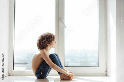 Topless kid portrait near the window. Sunny day outside. Bright white room. Boy poses, smiles and having fun alone. 