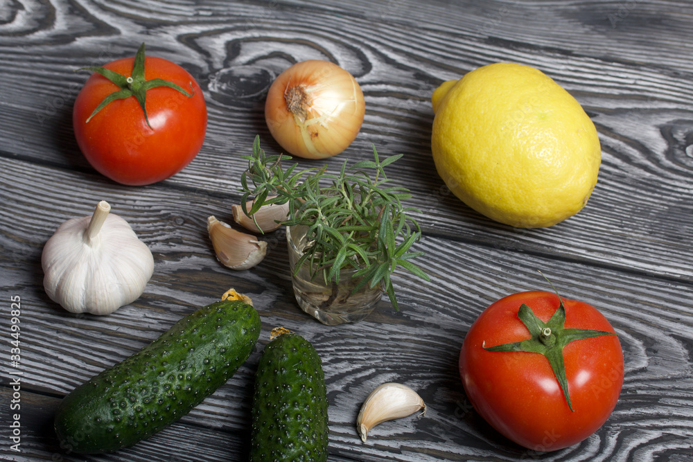 Tomatoes with green ponytails, onion, lemon and garlic head. Two green prickly cucumbers with yellow flowers. A glass with rosemary branches. They lie on the surface of brushed pine boards. Tomatoes with green ponytails, onion, lemon and garlic head. Two green prickly cucumbers with yellow flowers. A glass with rosemary branches. They lie on the surface of brushed pine boards.