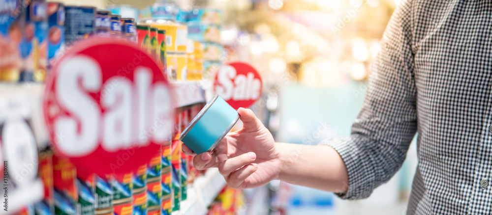 Fototapeta premium Asian man customer choosing canned food from shelf in supermarket or grocery store. Hoard instant food for quarantine at home during Coronavirus (COVID-19) pandemic. Consumerism concept