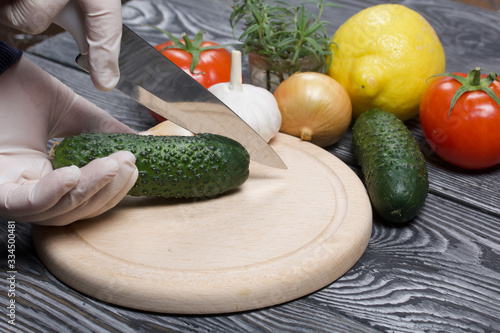 A man in rubber gloves cuts a cucumber with a knife on a cutting board. Nearby are red tomatoes with green ponytails, rosemary and garlic. On the surface of brushed pine boards.