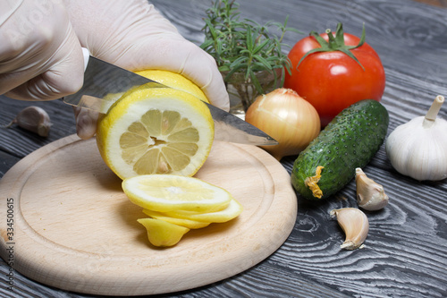 A man in rubber gloves cuts a lemon with a knife on a cutting board. Nearby are cucumber, tomato, rosemary and garlic. On the surface of brushed pine boards.