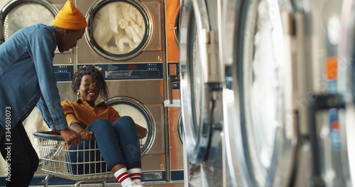Young African American cheerful couple having fun in laundry service room. Pretty happy girl sitting in carriage and guy riding her at working washing machines while cleaning clothes.