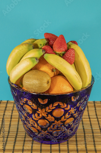 Fresh tropical and subtropical fruits in a large blue crystal vase. Wicker bamboo stand on the dining table, blue background photo