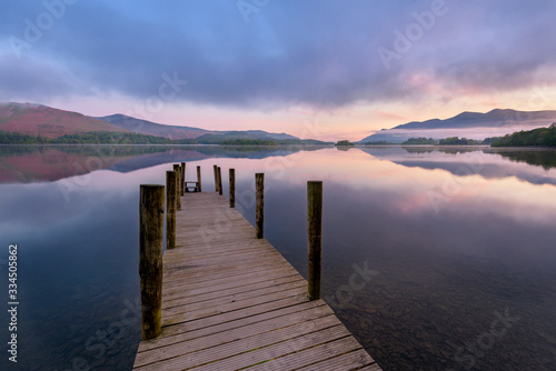 Still Reflections On A Calm Morning With Wooden Jetty In The Lake District, UK.