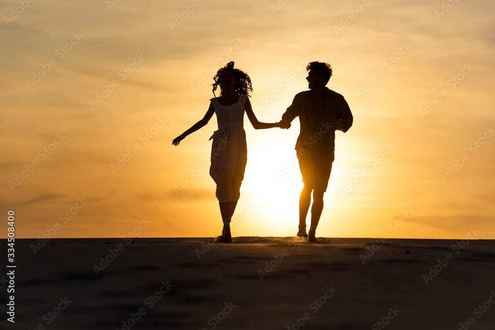 silhouettes of man and woman running on beach against sun during sunset