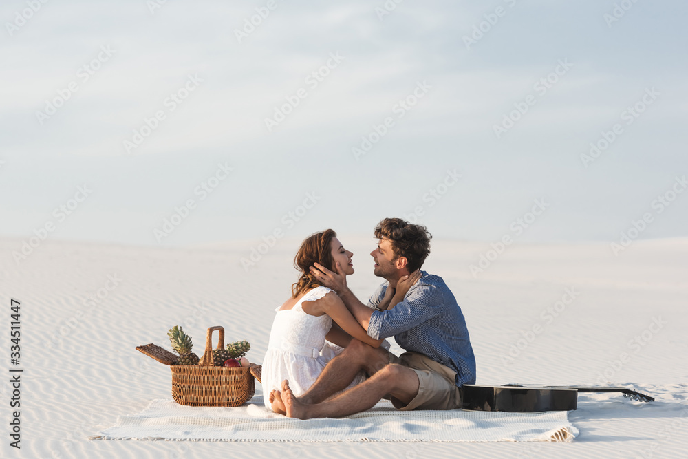 Fototapeta premium young couple hugging while sitting on blanket with basket of fruits and acoustic guitar on beach