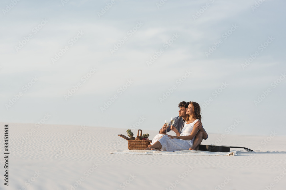 young couple drinking champagne while sitting on blanket with basket of fruits and acoustic guitar on beach