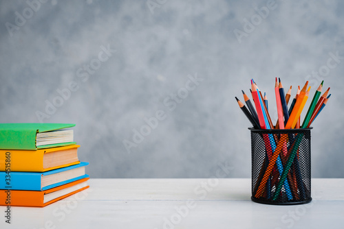 A stack of colorful textbooks and a glass of bright pencils on a gray- blue background.