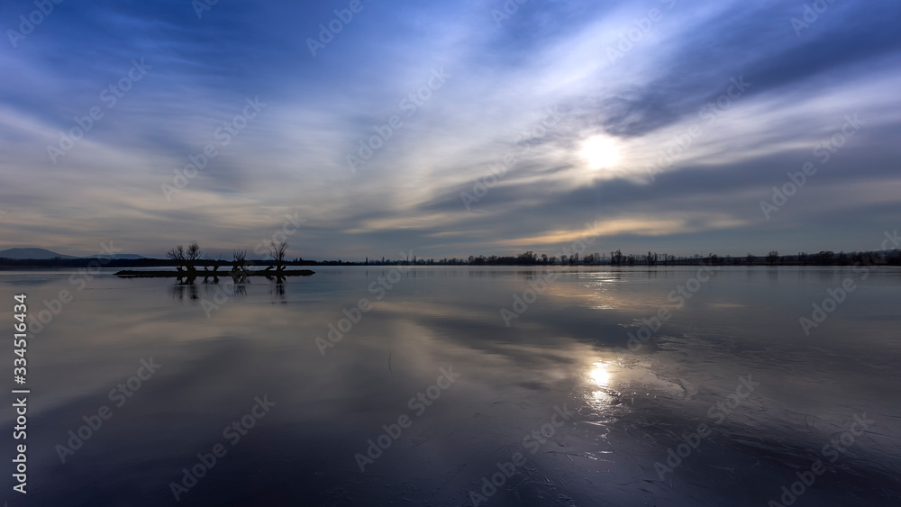 A stone island covered with willows in the middle of a pond by the setting sun with dramatic clouds A stone island covered with willows in the middle of a pond by the setting sun with dramatic clouds