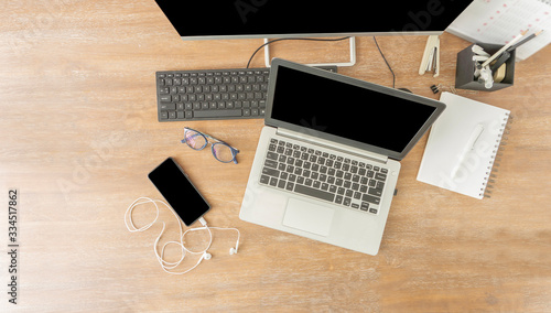 Top view of a wooden working desk including black screen laptop computer  keyboard  mobile cell phone  notebook  box of pens in an office